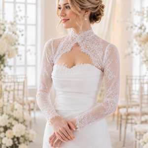 May include: A white lace bolero jacket featuring long sleeves and a high neckline with intricate floral patterns. The bolero is worn over a strapless white gown with a sweetheart neckline, showcasing a delicate scalloped edge along the lace trim.