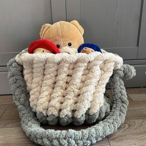 May include: A beige teddy bear peeks over the edge of a cream, gray, and white crocheted basket. Two smaller teddy bears wear a red and blue hat. The basket has a gray handle.