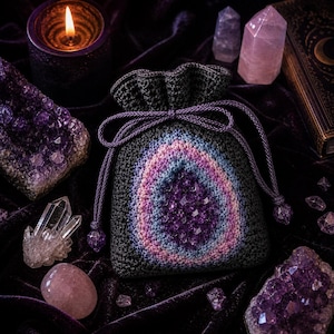Op de afbeelding: Een gehaakte trekkoordtas met een amethistgeode-ontwerp. De tas heeft een kleurengradatie van paars, blauw en roze. De afbeelding bevat ook amethistkristallen, een brandende kaars en andere kristallen.
