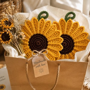 May include: Two round crochet sunflower pot holders with dark brown centers and bright yellow petals, each featuring a green loop. They are tucked into a brown paper gift bag with a tag reading Handmade with love and a small sun icon.