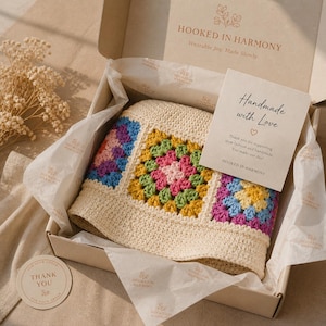 May include: A cream-colored crochet hat with colorful squares in a box. The squares feature pink, blue, green, and yellow. The box contains tissue paper and a card that says "Handmade with Love". The brand name "Hooked in Harmony" is visible.