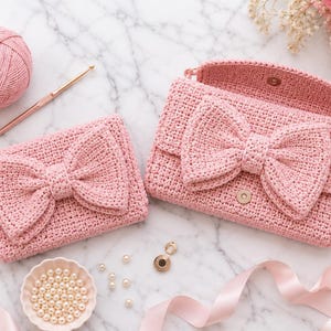 Puede incluir: Dos bolsos de ganchillo rosas con grandes lazos se muestran sobre una superficie de mármol. También hay una madeja de hilo rosa, un ganchillo y un cuenco pequeño con perlas. Una cinta y flores completan la escena.