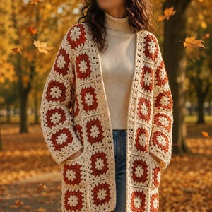 May include: Cream and rust-colored crochet cardigan with a floral pattern. The open-front sweater features a geometric design and pockets. The cardigan is worn over a white turtleneck and blue jeans, with autumn leaves in the background.