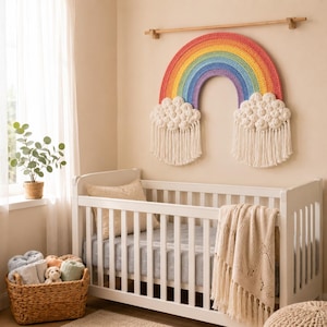 May include: A crocheted rainbow wall hanging with red, orange, yellow, green, blue, and purple bands, featuring white textured clouds at each end with long, hanging fringe, suspended from a wooden rod above a white wooden crib with bedding.