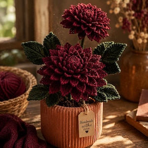May include: A handmade burgundy crochet flower arrangement in a terracotta pot. The flowers have detailed petals and green leaves. A small tag reads "Handmade with Love". The arrangement sits on a wooden surface, with a basket of yarn in the background.