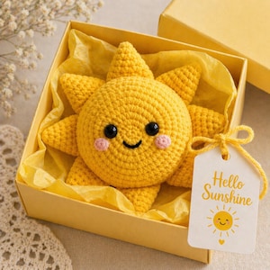May include: A handmade yellow crochet sun with a smiling face and pink cheeks, presented in a yellow gift box. The box contains yellow tissue paper and a tag that says "Hello Sunshine" with a sun illustration.