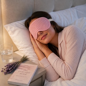 May include: A pink sleep mask with a white strap is worn by a person lying in bed. The mask is made of a knitted material. A book titled "the peaceful mind" and a glass of water are on the nightstand.