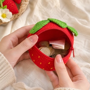 May include: A round, red crocheted coin purse shaped like a strawberry, featuring small yellow embroidered seeds and green crocheted leaves on top. The purse has a zippered opening containing several metallic coins and a folded paper banknote.