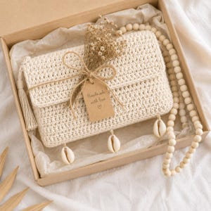 May include: A cream-colored, crocheted clutch with a flap closure, tassel, and seashell accents, presented in a cardboard box. The clutch is adorned with a tag that reads "Handmade with love." A dried flower bouquet and a beaded necklace complete the presentation.
