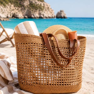 Puede incluir: Un bolso de playa marrón tejido con asas de cuero, lleno de una toalla a rayas, un sombrero de paja y un tubo de protector solar. El bolso está en una playa de arena.