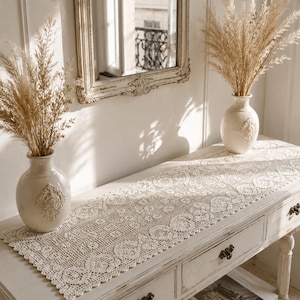 May include: A cream-colored, distressed wooden console table with a decorative, openwork lace runner. Two white vases with dried grasses sit on the table. A mirror with a distressed frame hangs on the wall behind the table.