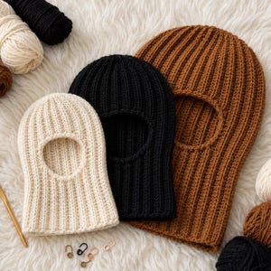 May include: Three knitted balaclavas in cream, black, and brown, with an oval opening for the face. The ribbed knit hats are displayed on a fluffy white surface, with yarn skeins and a crochet hook nearby. A cozy winter accessory.