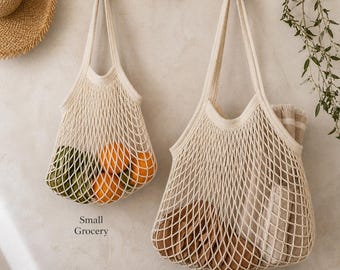 May include: Two off-white, open-weave cotton tote bags hanging on a wooden rack. One bag is labeled "Small Grocery" and contains green and orange fruit. The other bag, labeled "Large Beach", holds a folded striped towel and other items.