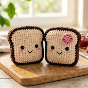 May include: Two crocheted toast slices with smiling faces and pink cheeks. One slice has a crocheted pink jam dollop. The toast has a light tan center and a dark brown crust. They are sitting on a wooden cutting board.