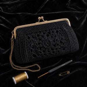 May include: A black crocheted clutch bag with a gold-coloured clasp and chain. The bag has an openwork pattern on the front. A spool of gold thread, a crochet hook, and a safety pin are also visible.