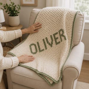 May include: A cream-coloured crocheted blanket featuring the name OLIVER in dark green block letters. The blanket has a textured stitch pattern and a dark green crocheted border along the edges, draped over a light-coloured upholstered chair.