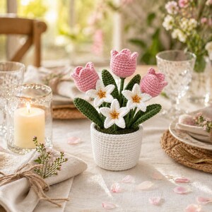 May include: A crocheted flower arrangement featuring pink tulips and white daffodils in a white pot. The handmade flowers have green stems and leaves. The arrangement is on a table with a lit candle and other decorative elements.