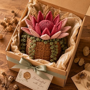 May include: A handcrafted crochet flower arrangement in a brown box. The arrangement features a large, pink and red flower with green foliage, presented on a bed of paper. A card with the words "Handcrafted with Love" is attached with a green ribbon.