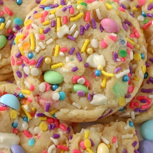 May include: Close-up of several soft-baked cookies covered in colorful sprinkles and pastel candies. The cookies have a light golden-brown color and are partially broken, revealing a soft interior. The sprinkles are in various colors, including yellow, pink, purple, and blue.