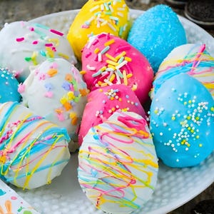 May include: A plate of colorful, decorated Easter egg treats. The eggs are coated in white, pink, yellow, and blue frosting, with sprinkles and drizzled icing. Some eggs have colorful drizzle patterns, while others are covered in sprinkles.