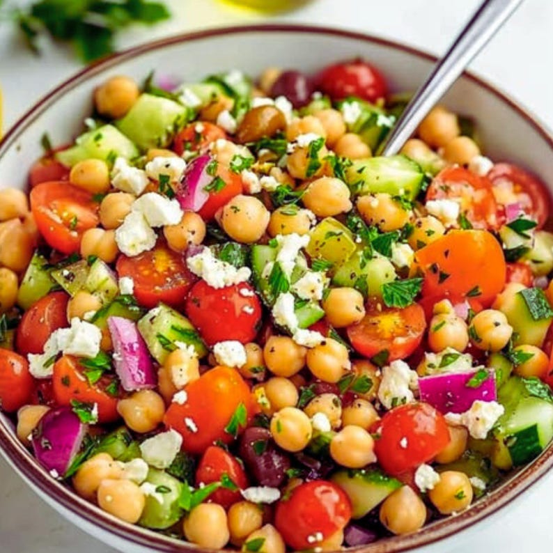 May include: A vibrant salad in a white bowl, featuring a mix of ingredients. The salad includes halved red cherry tomatoes, diced cucumbers, chickpeas, red onion, olives, and crumbled white cheese, garnished with fresh green herbs. A silver spoon rests in the salad.