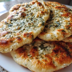 May include: A close-up of several golden-brown, flatbreads topped with a generous scattering of fresh green herbs. The bread has a slightly charred appearance, suggesting it was baked at high heat. The bread is on a white plate.