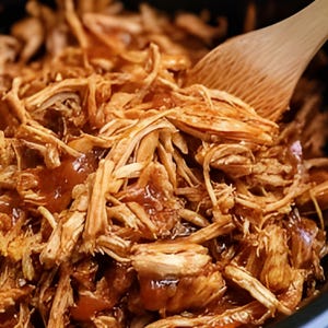 Pode incluir: Close-up de carne de porco desfiada com um molho castanho brilhante. A carne desfiada é de cor castanho claro, e uma colher de pau é parcialmente visível, mexendo o prato. A comida parece recém-cozinhada e pronta para servir.