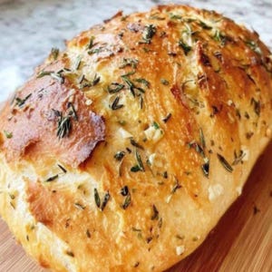 May include: A golden-brown loaf of bread, sprinkled with fresh green herbs and visible garlic pieces. The bread has a rustic appearance with a slightly cracked crust. The image showcases a freshly baked, appetizing loaf.