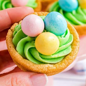 May include: A close-up of a mini tart with green frosting and three colorful candies. The tart shell is golden brown, and the frosting is swirled to resemble a nest. The candies are pink, blue, and yellow.