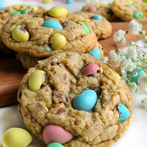 May include: Close-up of several Easter cookies with colorful candy-coated chocolate eggs. The cookies are golden brown and have a slightly textured surface. Some of the eggs are yellow, blue, and pink. The cookies are arranged on a wooden board.