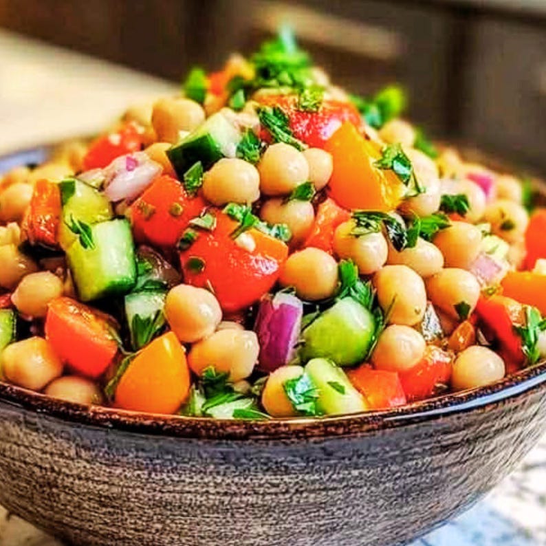May include: A colorful salad in a textured bowl. The salad features chickpeas, diced tomatoes, cucumbers, red onion, and fresh herbs. The tomatoes are red and orange, the cucumbers are green, and the onion is purple. The bowl is a neutral brown color.