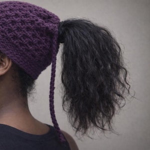 May include: A dark purple, crocheted hat with a long, plaited tail. The hat covers the top of the head and part of the ear. Black hair is pulled through the hat and styled into a ponytail.