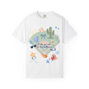 May include: White t-shirt with a baseball-themed graphic. The design includes a baseball field, baseball, cactus, sunglasses, hot dog, popcorn, and the text "Take me out to the ballgame."
