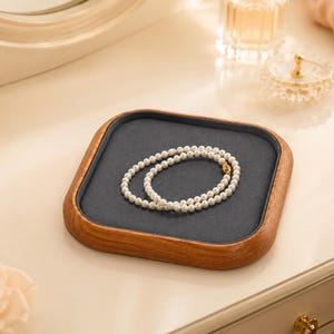 May include: A square wooden jewelry tray with a dark gray felt lining, featuring a single strand of white pearl beads with a small gold-toned clasp resting in the center. The tray has rounded corners and a natural wood grain finish.