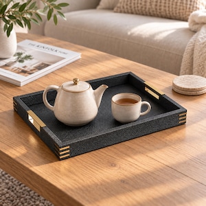 Puede incluir: Una bandeja rectangular negra con superficie texturizada, asas de metal dorado y detalles dorados en las esquinas. Una tetera de cerámica color crema con motas y un pomo dorado en la tapa, junto a una taza a juego, descansan sobre la bandeja.