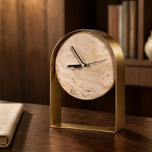 Puede incluir: Un reloj de mesa circular con una esfera de piedra veteada en tonos beige y bronce con un patrón natural de remolinos. El reloj está encajado en un marco de metal dorado mate con una parte superior arqueada y base rectangular, con manecillas negras.