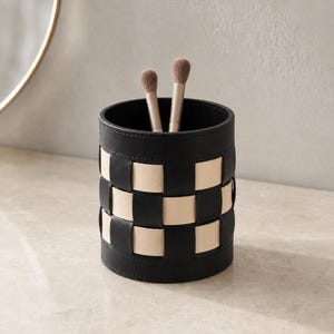May include: A cylindrical container featuring a woven pattern of black and light beige strips. The interior is lined in black, and two makeup brushes with light brown bristles and metallic handles are placed upright inside the holder.
