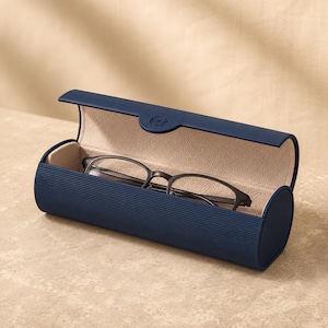 May include: A navy blue, cylindrical, hard-shell glasses case with a textured, ribbed exterior and a smooth, beige-colored interior lining. The case is open, revealing a pair of dark-rimmed eyeglasses resting inside the protective compartment.