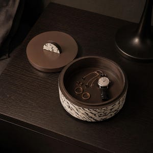 Puede incluir: Una caja de almacenamiento redonda de color marrón con tapa extraíble que presenta un patrón moteado en blanco y negro en el exterior. La tapa tiene un asa semicircular con un acabado moteado a juego. Dentro hay dos anillos, una cadena y un reloj.