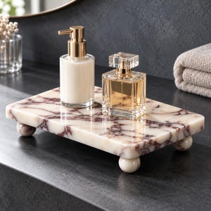 Puede incluir: Una bandeja rectangular de mármol blanco y burdeos con cuatro pies esféricos. La bandeja sostiene un dispensador de jabón de vidrio transparente con bomba dorada y líquido blanco, y un frasco de perfume cuadrado de vidrio con tapa dorada.