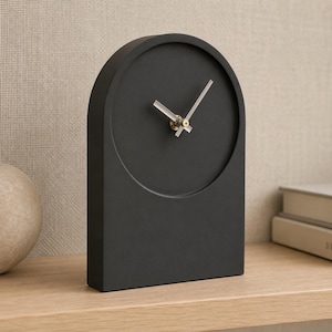 May include: A matte black tabletop clock with an arched top silhouette. The clock face is circular and recessed into the frame, featuring two simple silver-toned hands at the center. The design is solid, smooth, and lacks any numerical markings.