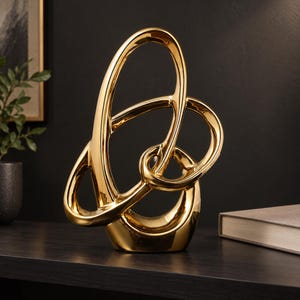 Peut inclure: Une sculpture en céramique dorée présentant un design en boucle nouée, fluide et abstrait. La surface est hautement réfléchissante et lisse, créant un aspect métallique poli avec des espaces ouverts interconnectés dans toute la structure.