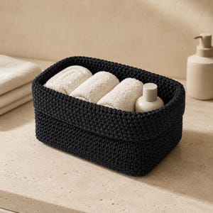 May include: A rectangular black woven storage basket containing three rolled beige towels and a small beige bottle with a cylindrical cap. The basket features a thick, textured knit construction with a folded top edge, providing a structured container.