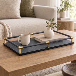 May include: A rectangular black tray with a textured surface, featuring a raised railing wrapped in dark material and accented with gold-toned metal corner posts and supports. A beige ceramic mug and a small white vase with greenery sit on the tray.