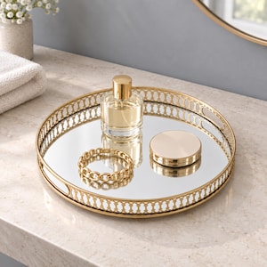 Puede incluir: Una bandeja decorativa redonda con marco de metal dorado con patrón de celosía y base de espejo. La bandeja contiene un frasco de perfume de vidrio transparente con tapa dorada, una pulsera de eslabones dorados y un pequeño estuche compacto.