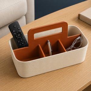 Può includere: Un contenitore rettangolare con esterno color crema dal sottile motivo a spina di pesce e interno marrone chiaro a contrasto. L'organizer presenta una maniglia centrale e tre scomparti interni che contengono un telecomando, una penna e occhiali.