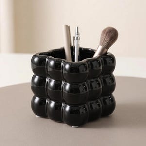 Könnte beinhalten: Ein glänzender schwarzer Schreibtisch-Organizer aus Keramik mit einer dreidimensionalen, noppenartigen Oberflächenstruktur. Der quadratische Behälter enthält zwei Stifte und einen Make-up-Pinsel mit braunen Borsten und steht auf kleinen Füßen.