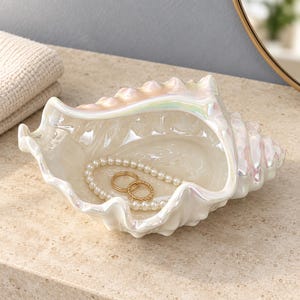 Peut inclure: Un plat en forme de coquille de conque en céramique blanche avec une finition irisée. À l'intérieur de la coquille se trouvent un collier de perles blanches et deux bagues en métal doré. La coquille présente un extérieur texturé et spiralé.