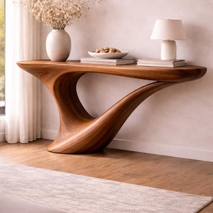 Puede incluir: Una consola de madera moderna y escultural con un diseño curvo. La mesa está hecha de madera de tonos cálidos y presenta un jarrón con flores secas, una lámpara pequeña y un cuenco con nueces. La mesa mide 183 cm de largo.