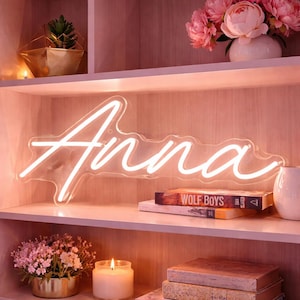 Può includere: Un'insegna al neon bianca con il nome "Anna" scritto in corsivo. L'insegna è posta su una mensola con libri, fiori e una candela, creando un'atmosfera accogliente e decorativa.